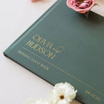 Green wedding guest book with 'Olivia & Hudson' on a light surface with flowers.