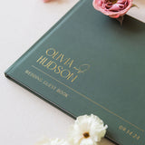 Green wedding guest book with 'Olivia & Hudson' on a light surface with flowers.