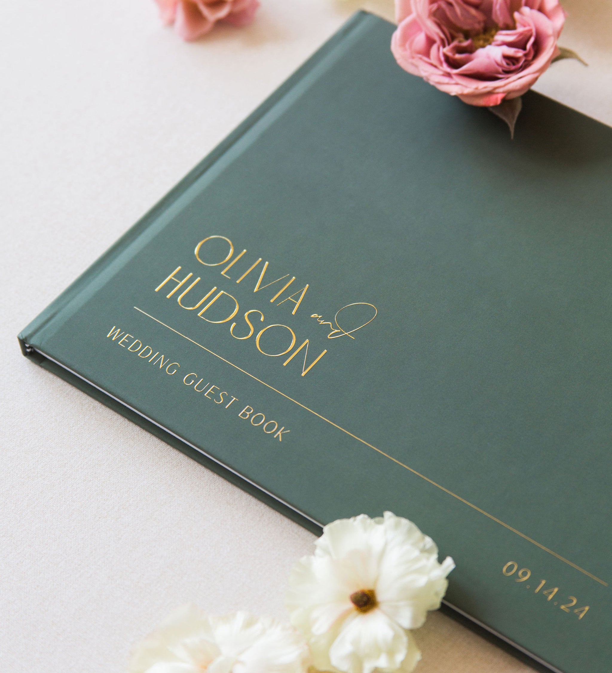 Green wedding guest book with 'Olivia & Hudson' on a light surface with flowers.
