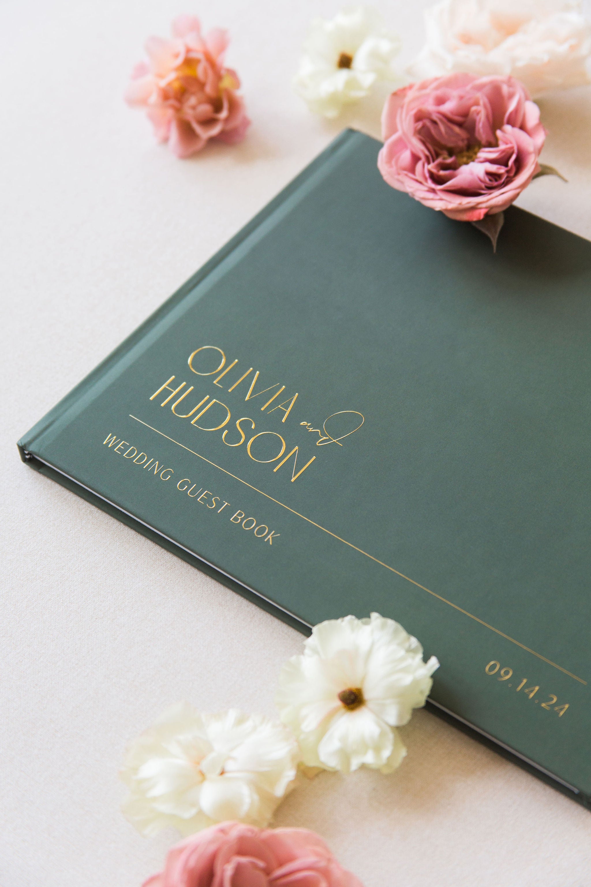 Green wedding guest book with 'Olivia & Hudson' on a light surface with flowers.
